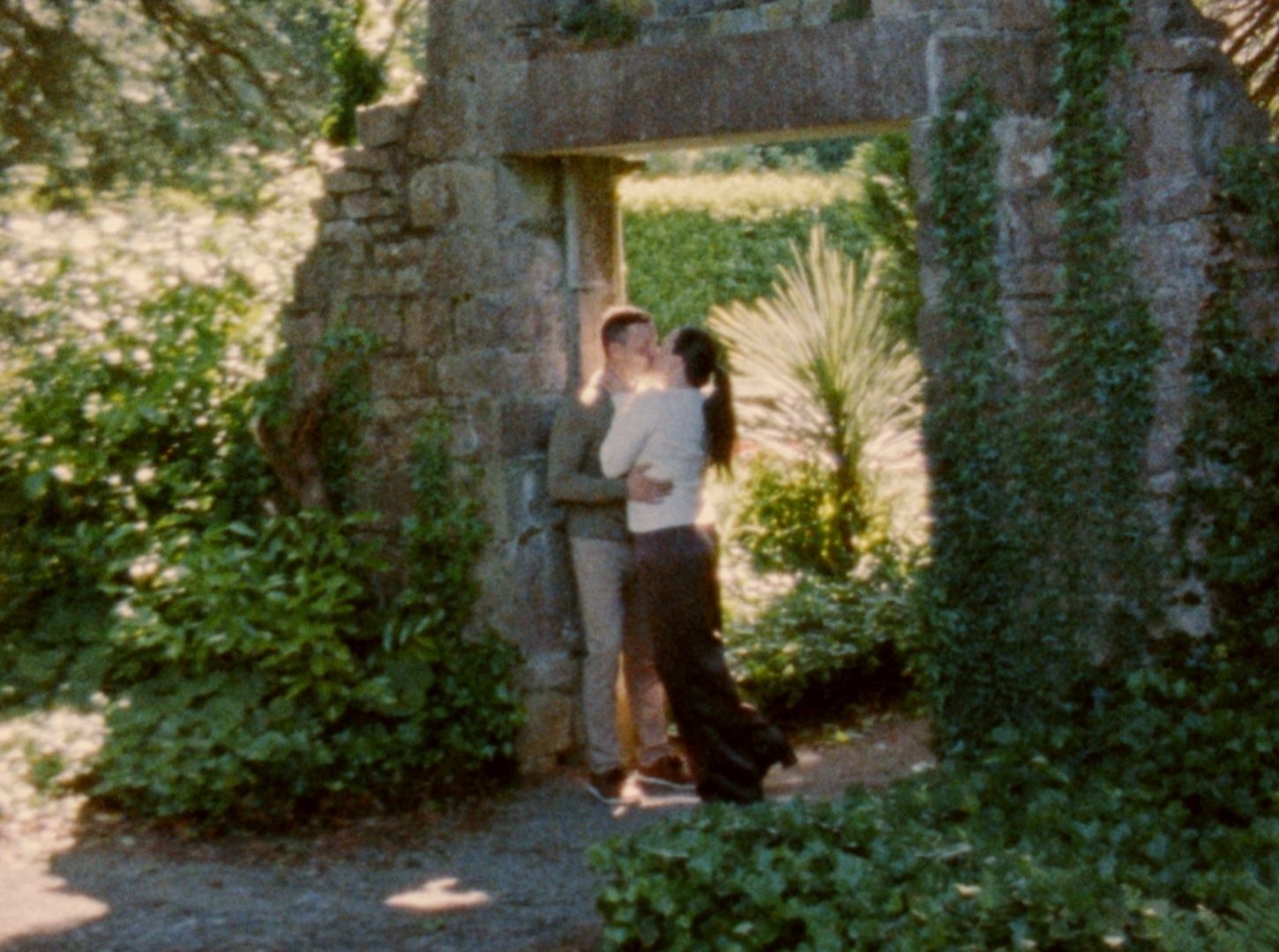 Couple embracing in a romantic stone garden archway surrounded by lush green foliage and plants.