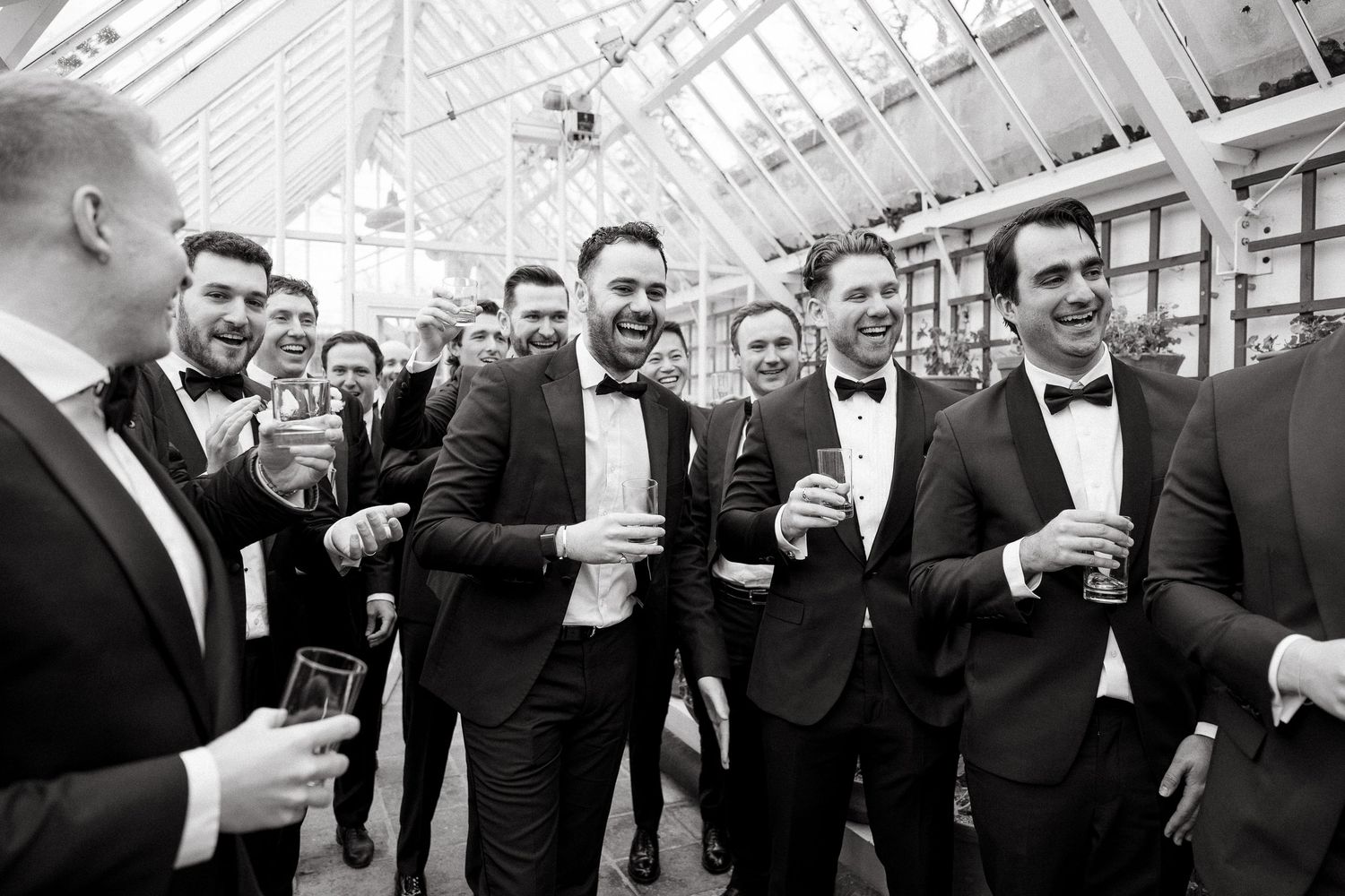 A group of groomsmen in black tuxedos raise their glasses in celebration at a wedding event.