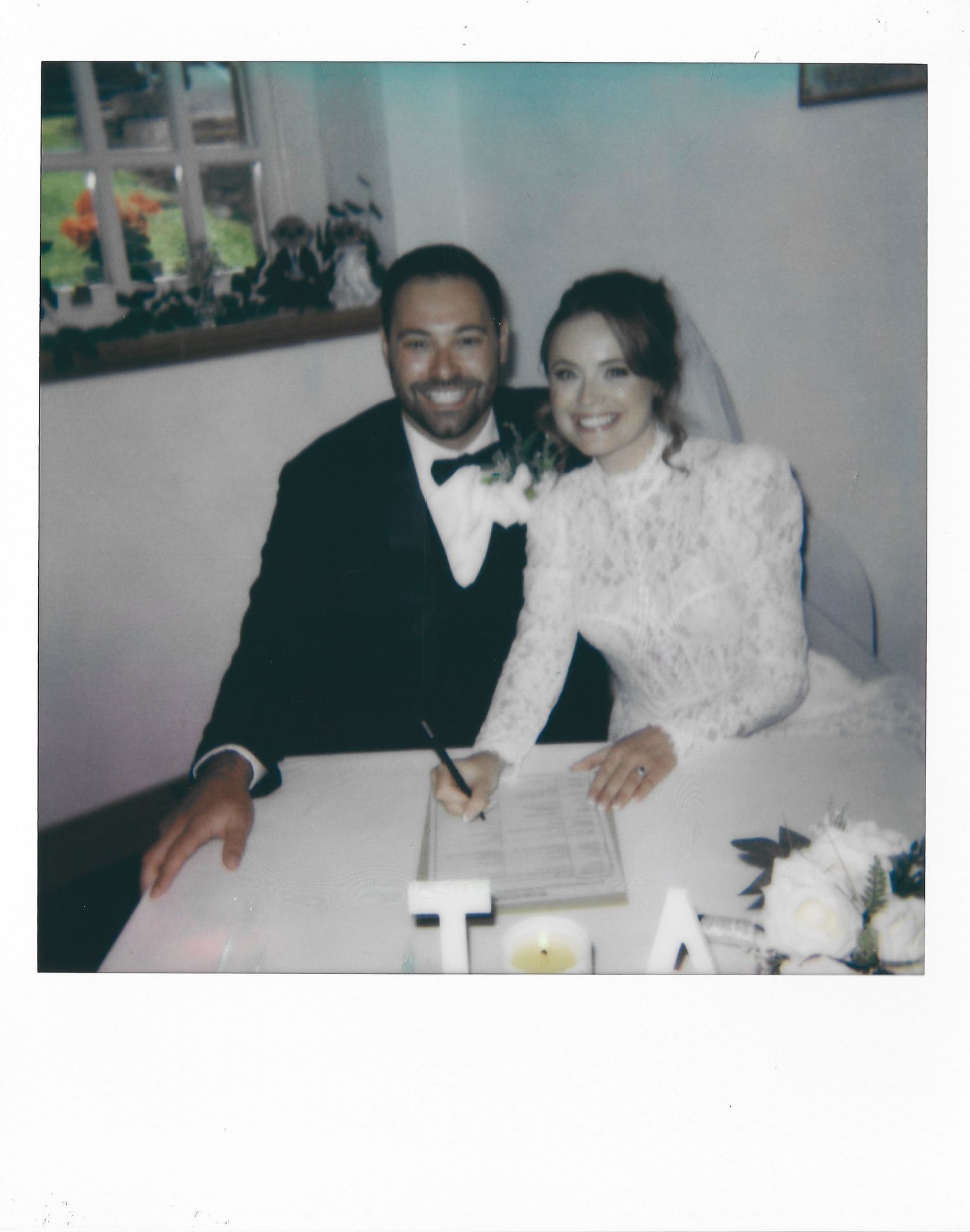 A couple in formal wedding attire sit together at a reception table signing documents.