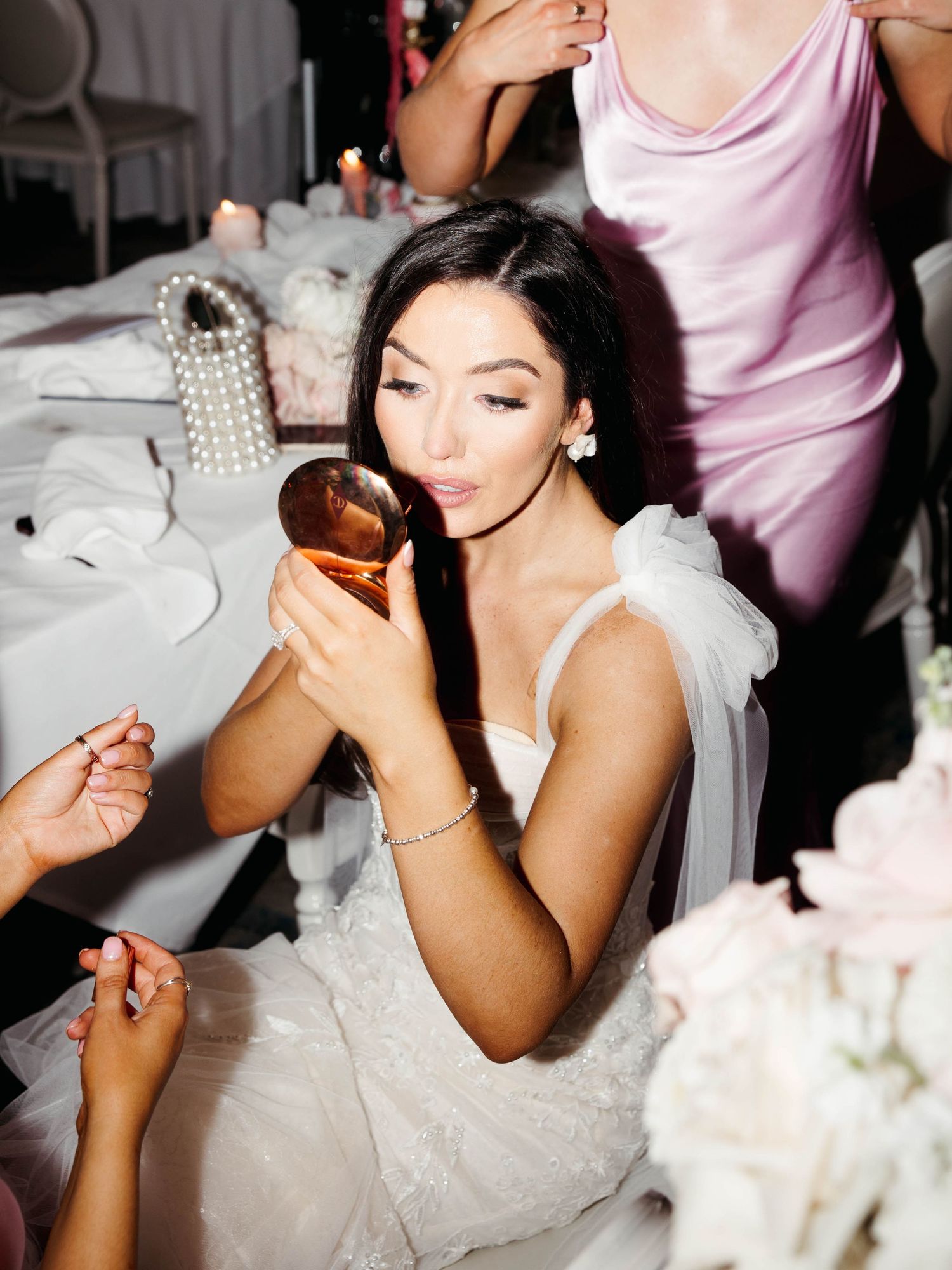 A person in a white dress holds a compact mirror while getting ready at a table with white floral decorations.
