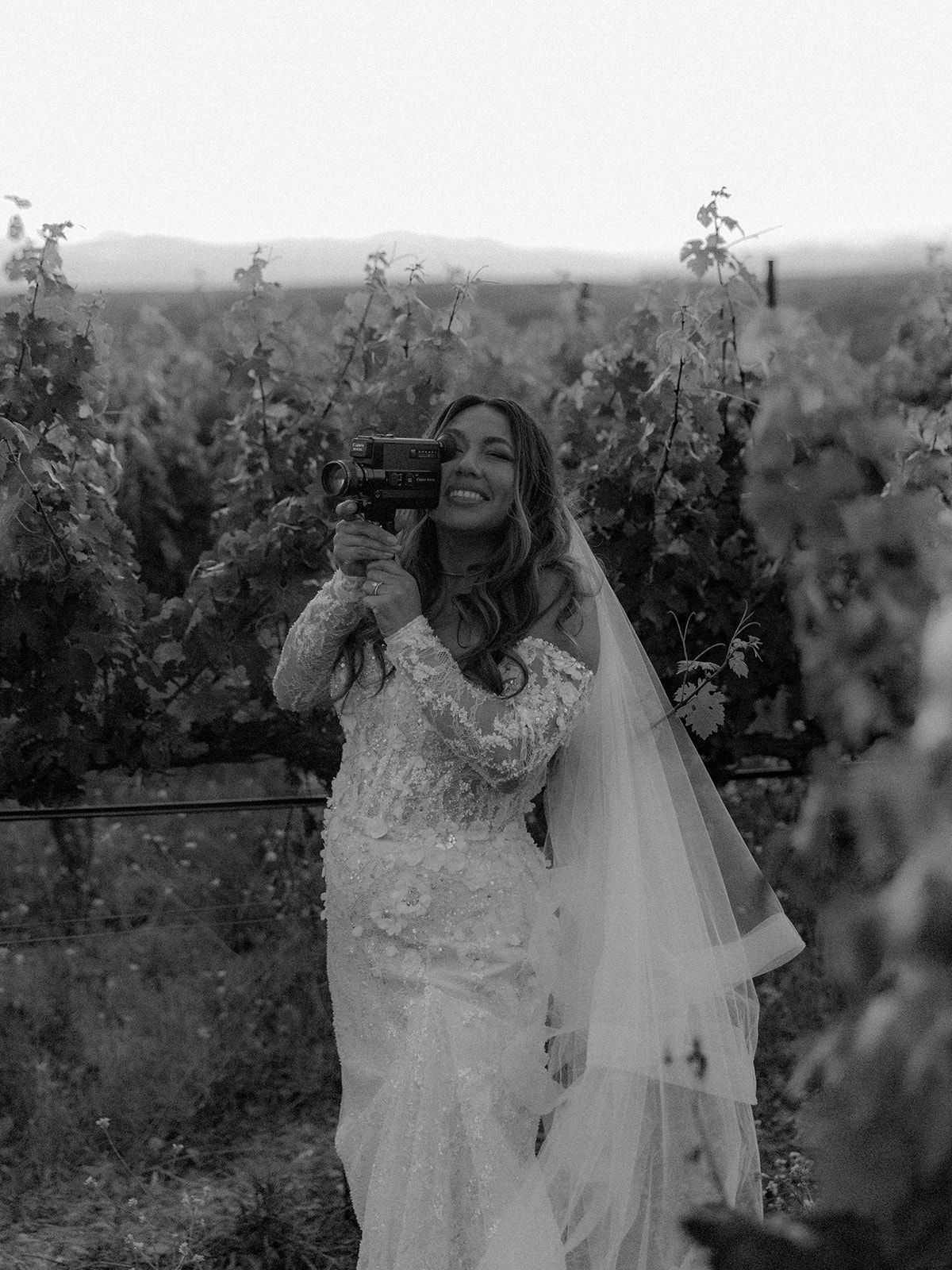 A bride in a lace wedding dress raises a wine glass while standing in a vineyard, captured in black and white.