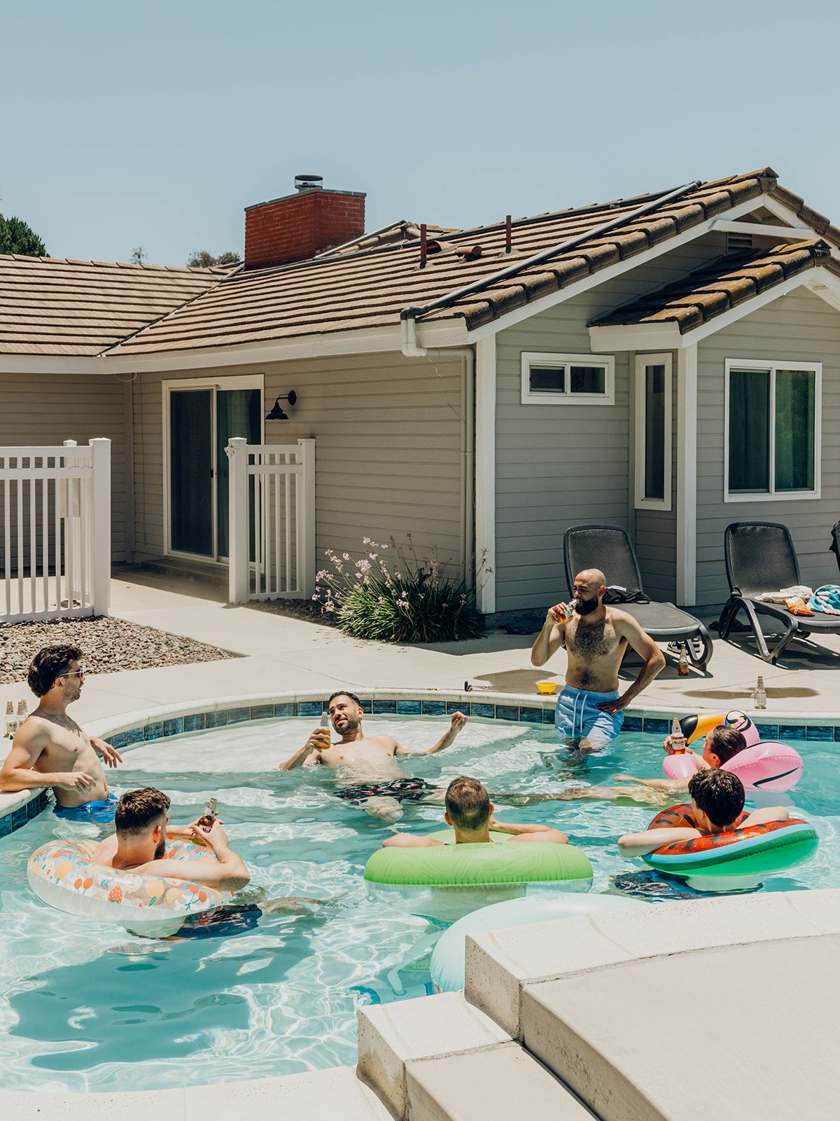 People enjoying a summer pool party with floaties in the backyard of a tan and grey house with a pool.