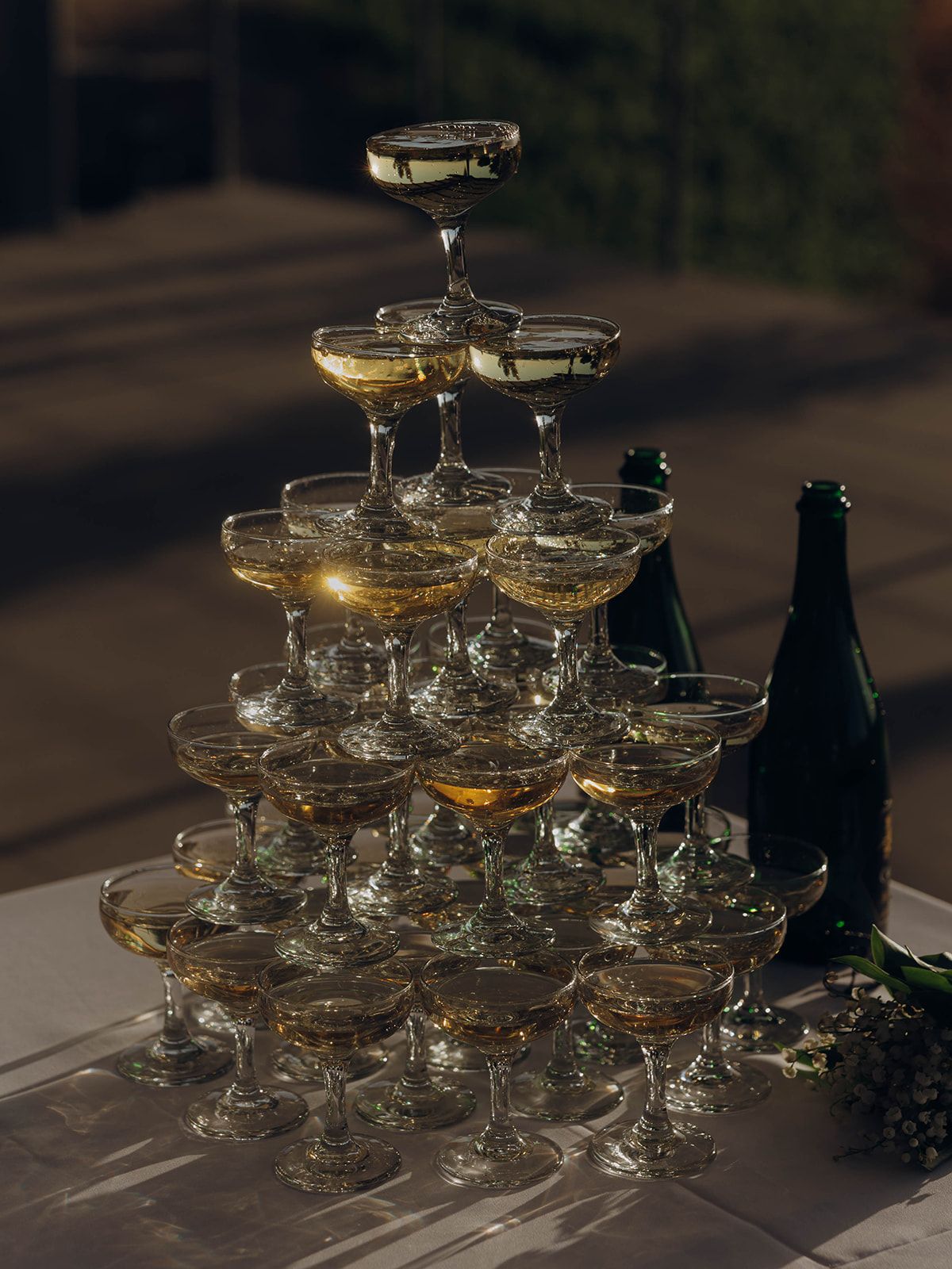 A champagne tower stacks elegantly filled coupe glasses with golden bubbling liquid beside dark bottles at dusk.