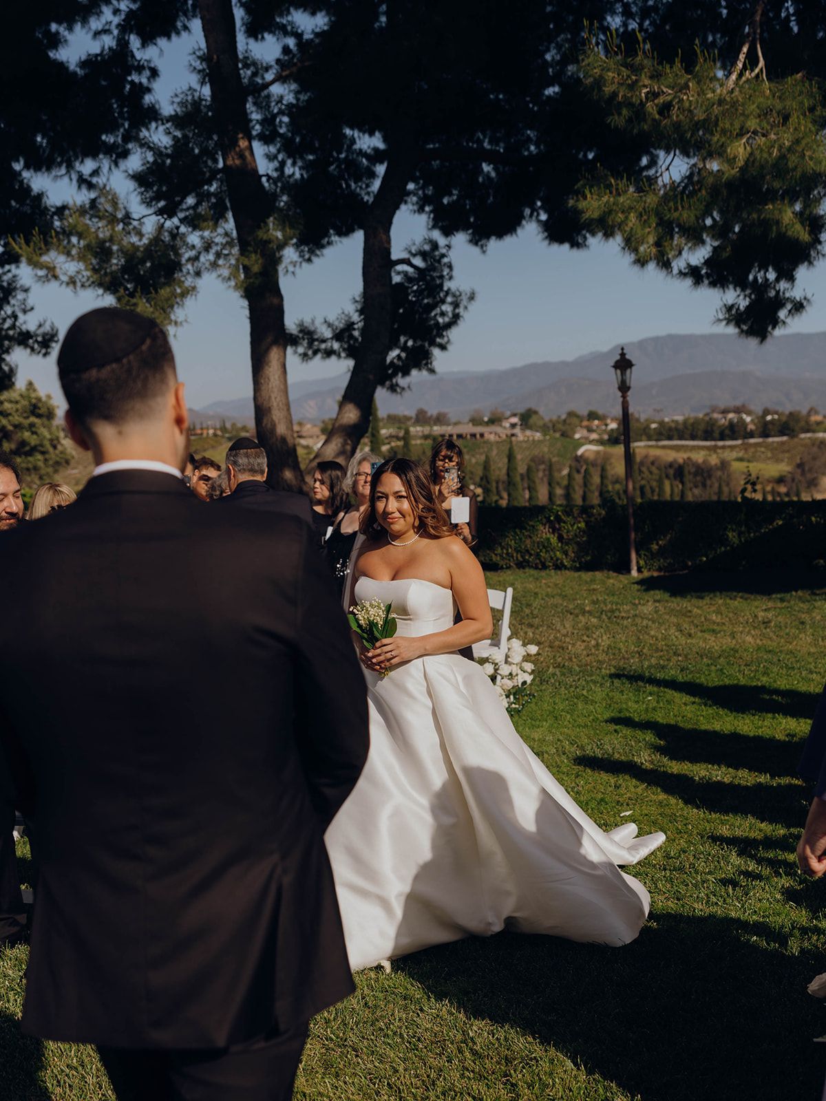 Outdoor wedding ceremony takes place on grassy lawn with mountain views in background and elegant flowing white gown.