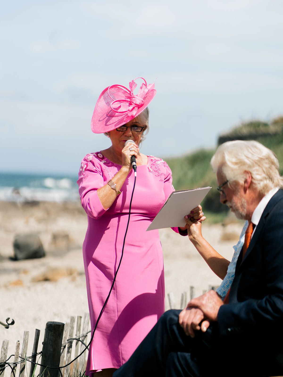 A woman in bright pink dress and hat speaks at outdoor event near the ocean.