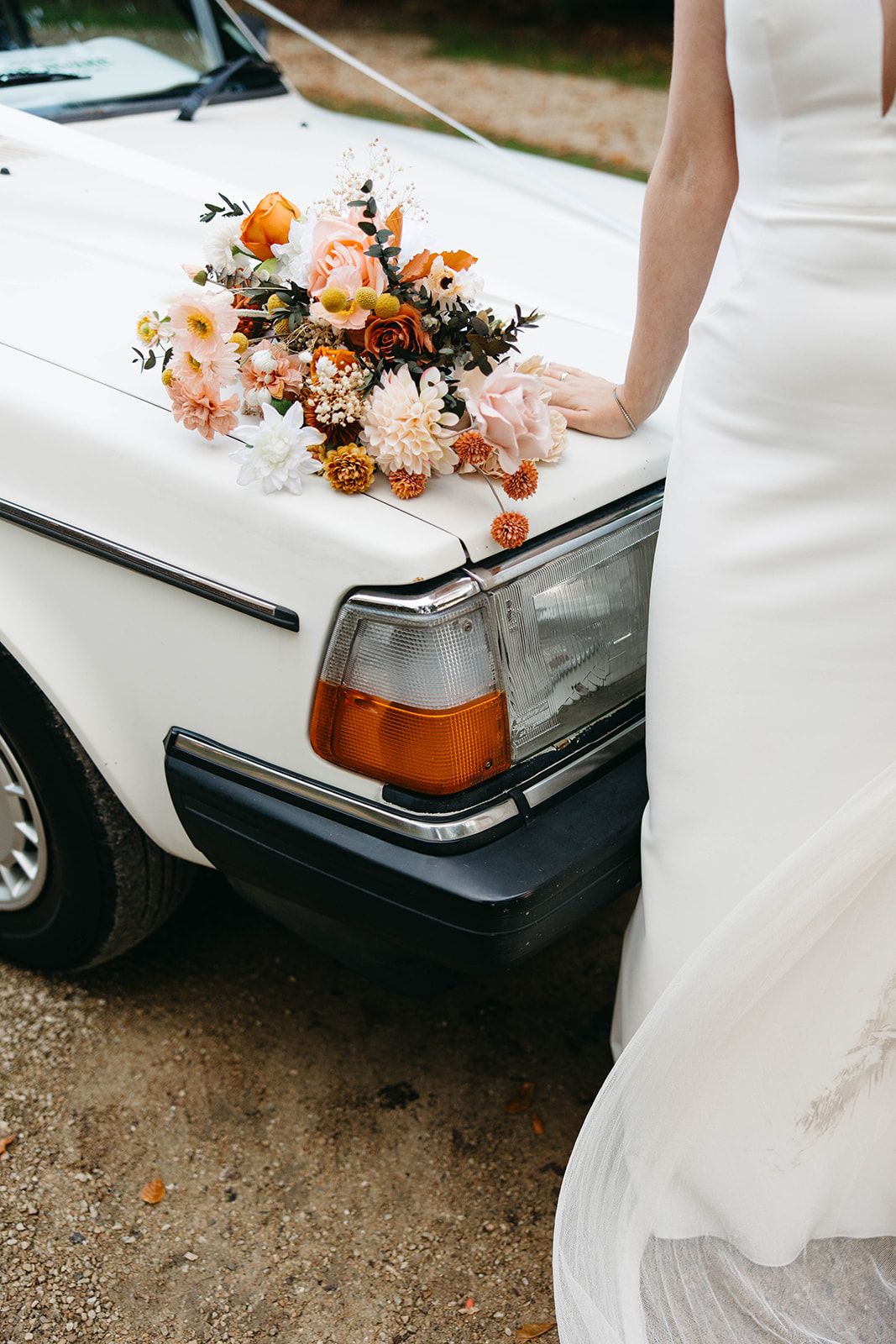 A vintage white car decorated with a peach and orange floral bouquet sits next to a flowing white wedding dress.