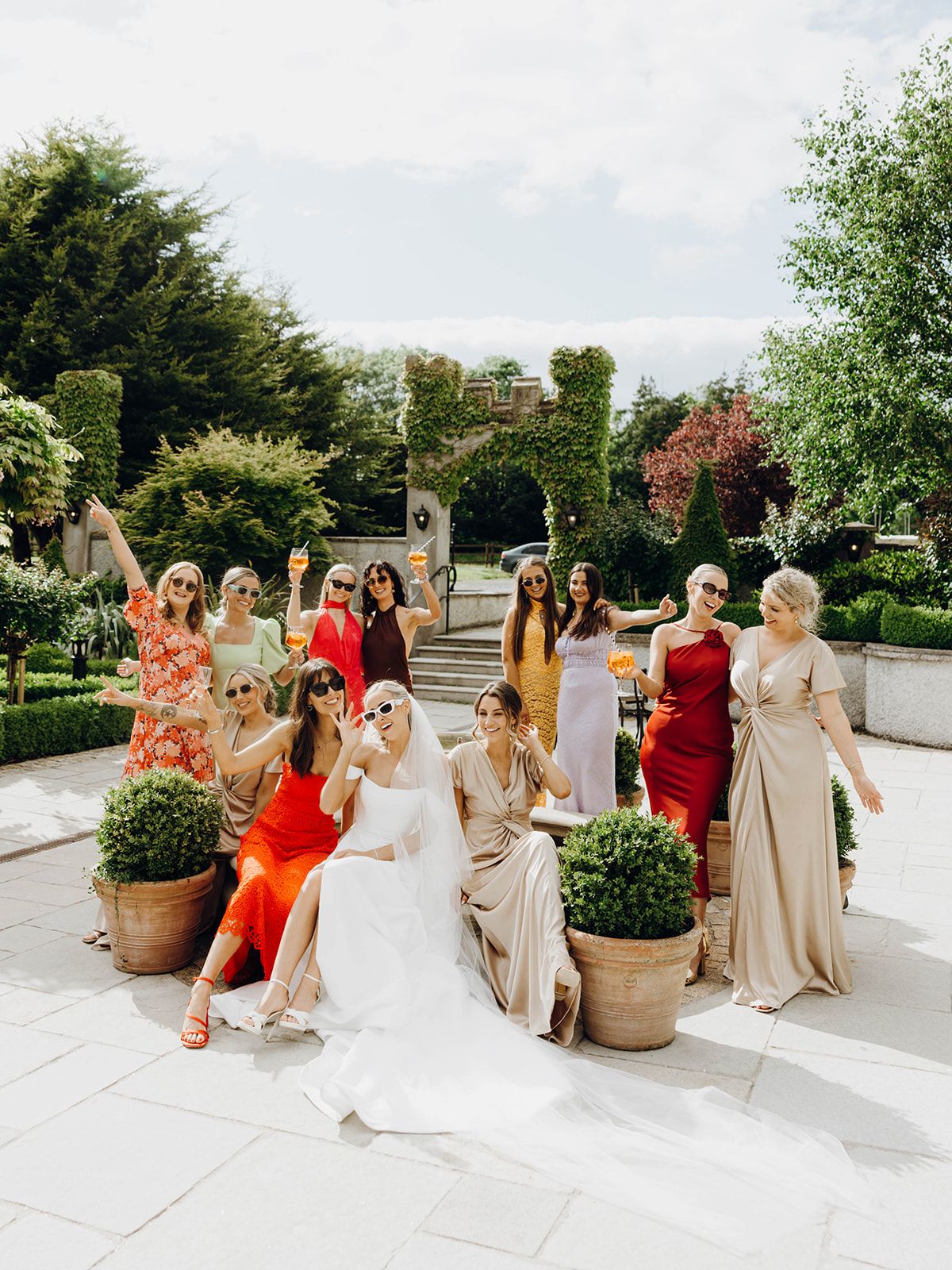 A group in formal attire poses together at an outdoor garden wedding with greenery and stone stairs in the background.