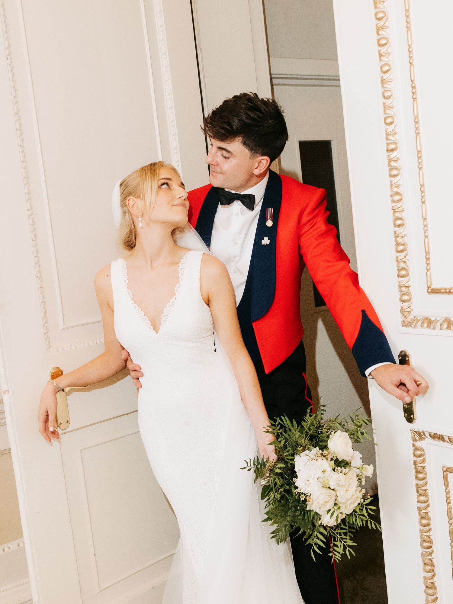 A couple in wedding attire pose together against an ornate door, with the groom in a red jacket and bride holding a floral bouquet.