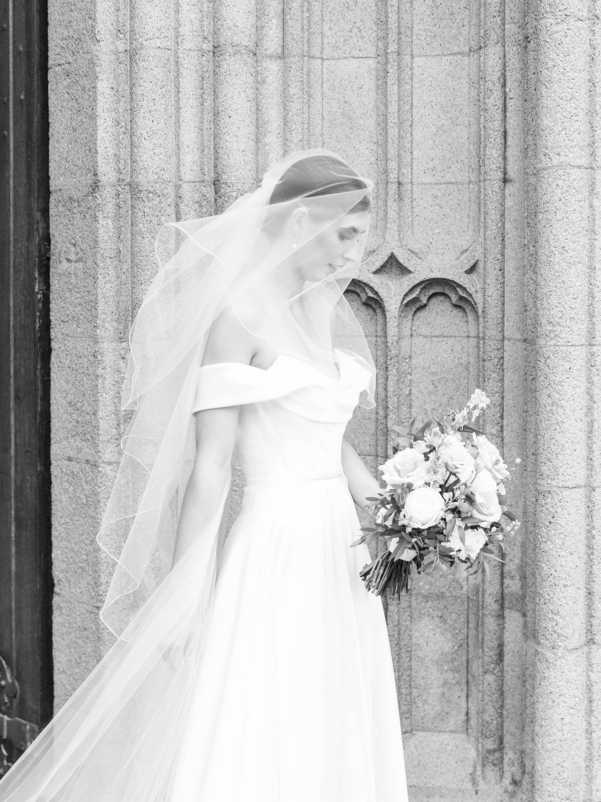 An elegant bride in a classic white gown and long veil poses beside a stone church column.