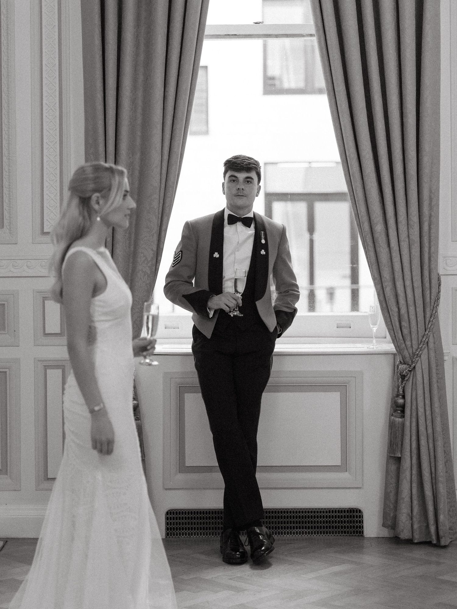 Wedding couple in elegant attire stand in ornate room near large windows and drapes in dramatic black and white photo.
