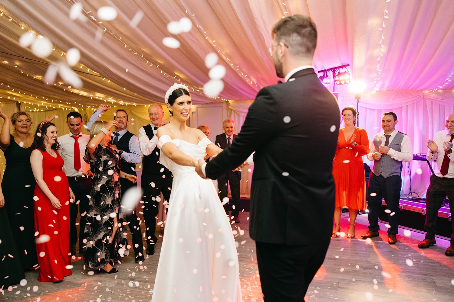 Newlyweds share their first dance as confetti falls on a pink-lit ballroom dance floor surrounded by wedding guests.