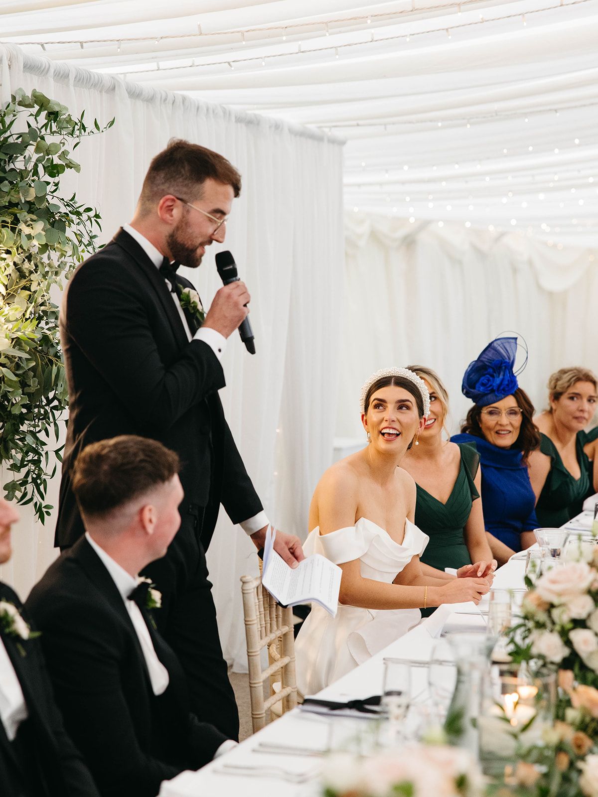 A wedding reception speech moment at an elegant white-draped venue with floral decor and seated guests in formal attire.