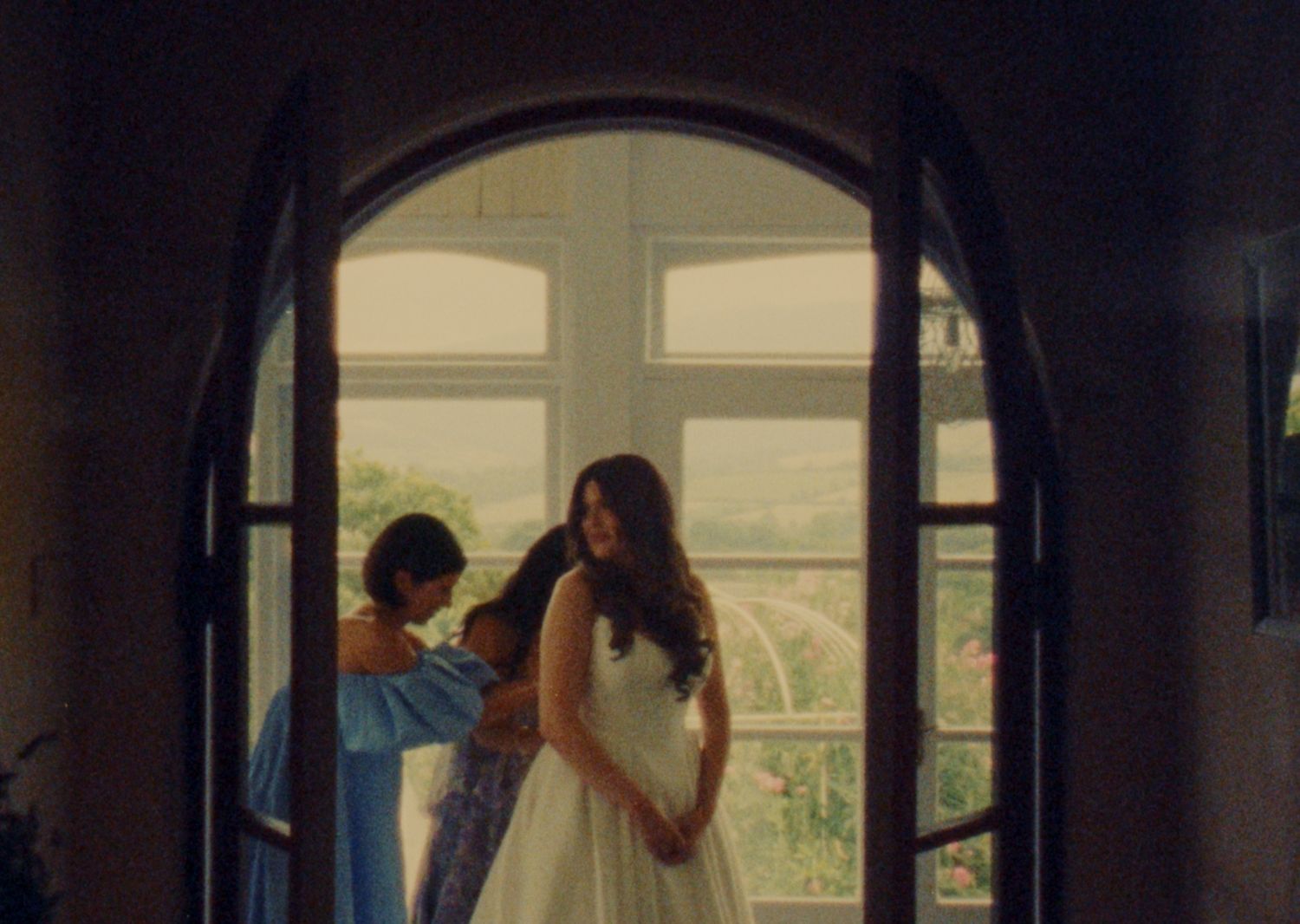 Bride in white gown stands by arched window as bridesmaid in blue dress assists her during wedding preparations.