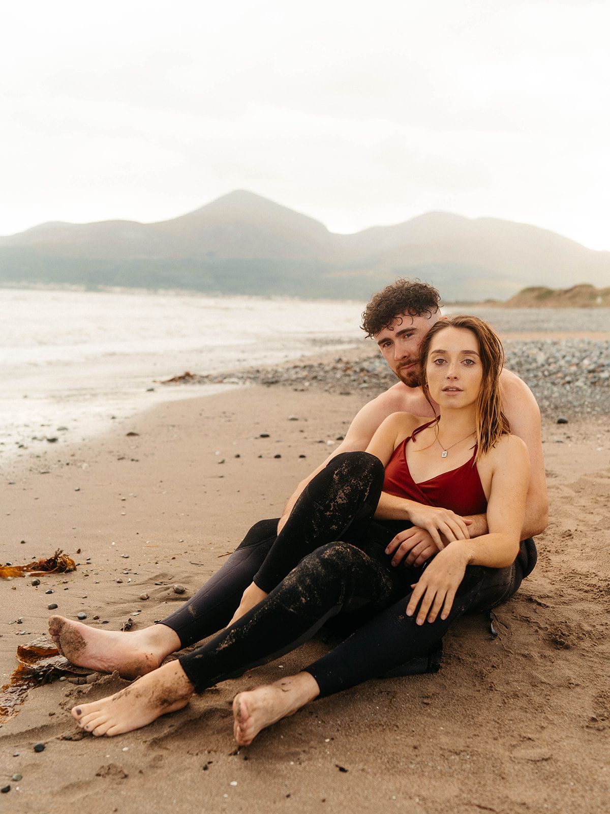 A couple sits together on a sandy beach, mountains in the background, wearing black leggings and a red top.