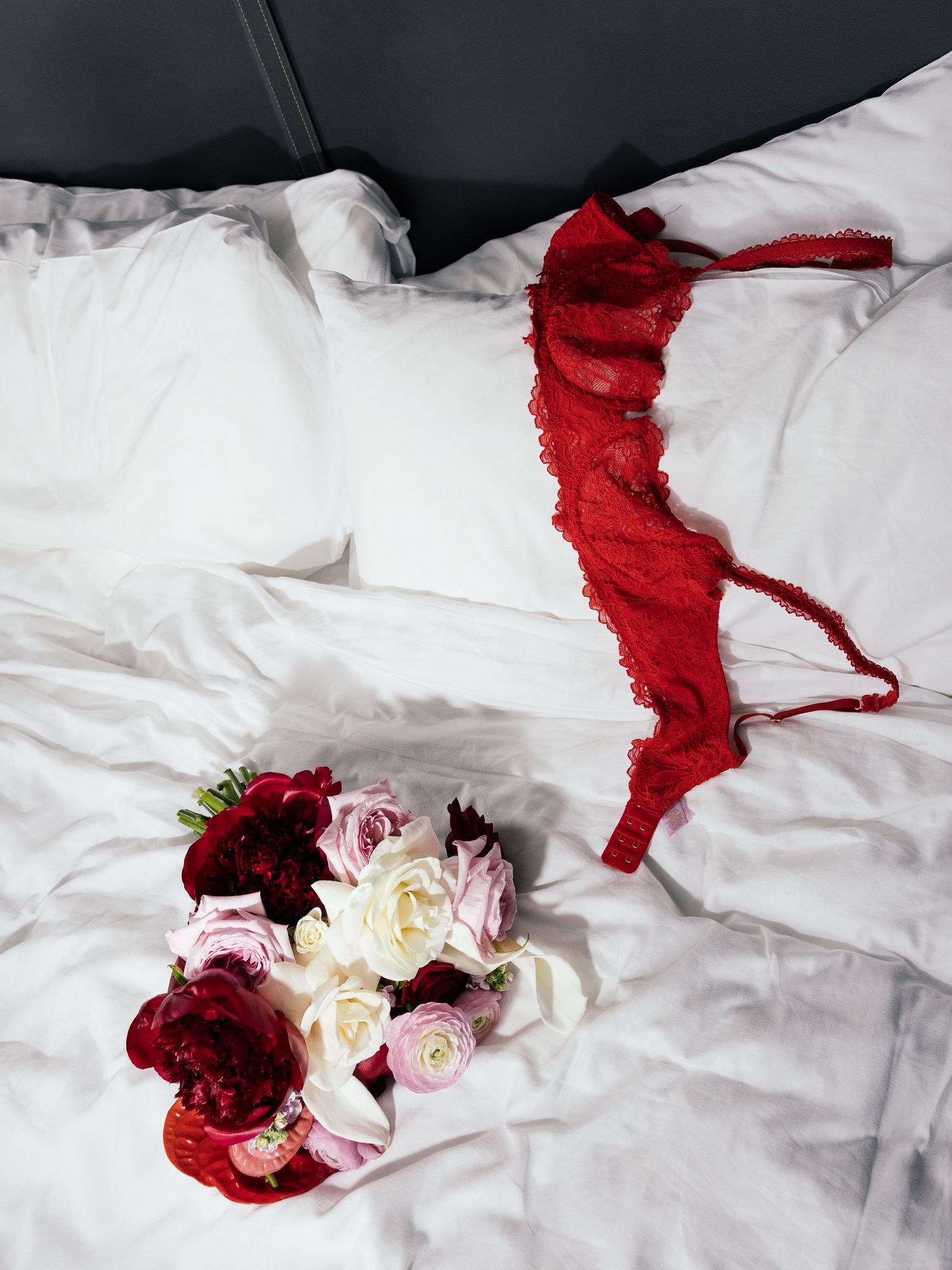 Red lace lingerie set and romantic bouquet of roses on white bed sheets.