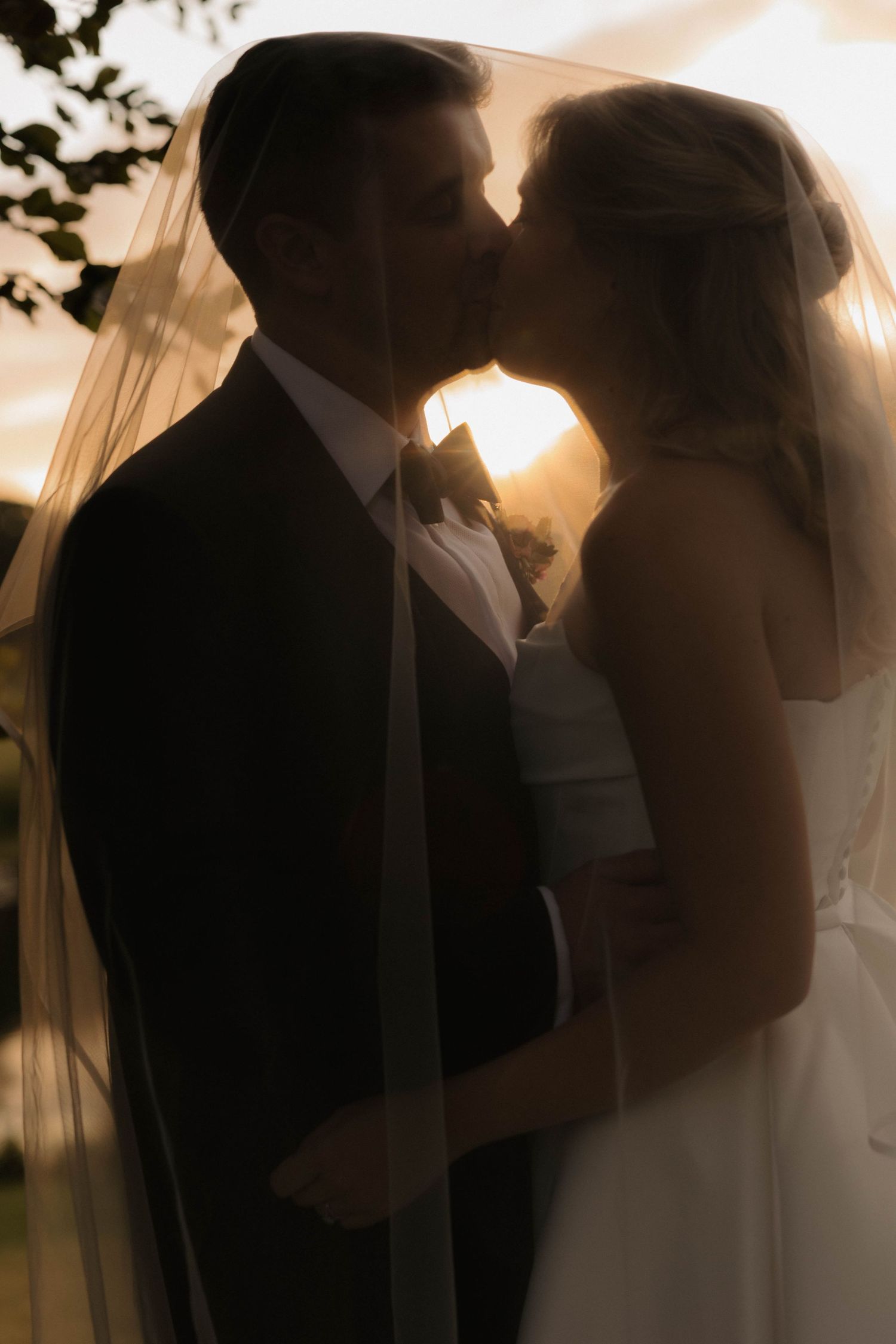 Silhouetted couple sharing intimate moments at sunset with wedding veil flowing.