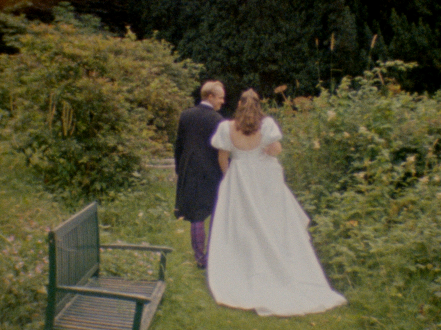 A couple in wedding attire poses in a garden setting with lush greenery and flowers in the background.
