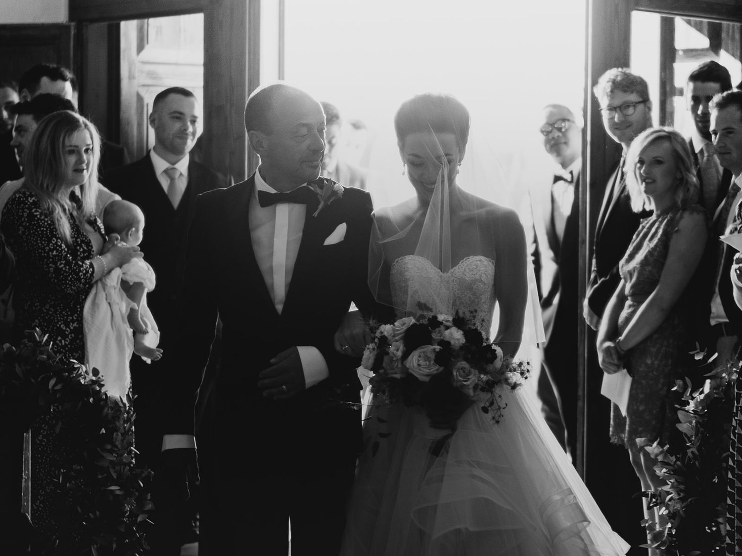 A black and white wedding ceremony photo showing a couple walking down the aisle surrounded by guests.