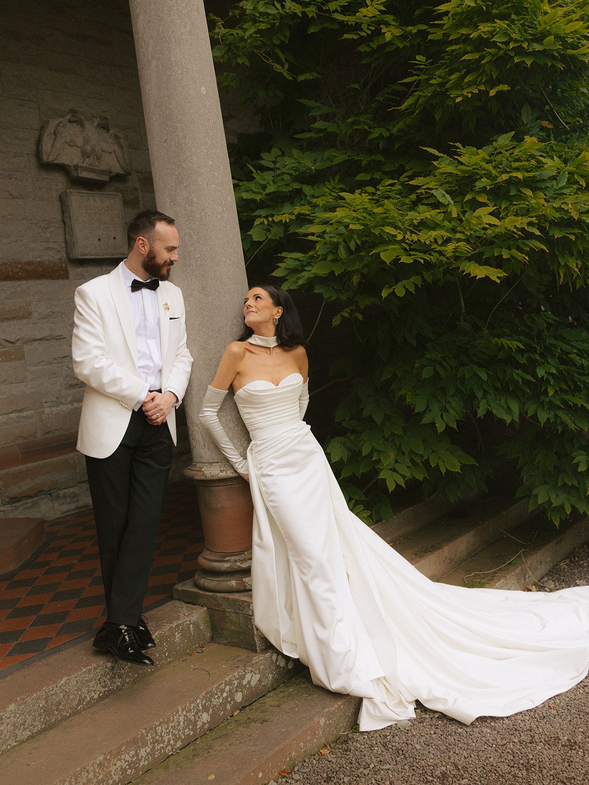 A romantic wedding couple poses together in a garden setting with stone columns and lush greenery in the background.
