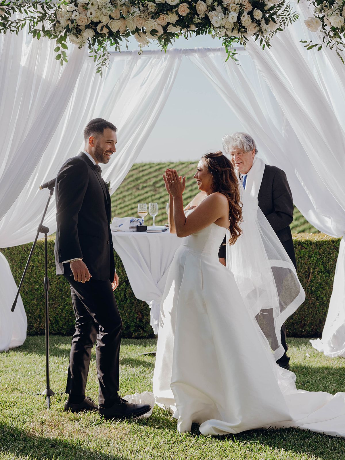 Wedding ceremony under a white draped arch with floral decor in an outdoor vineyard setting.