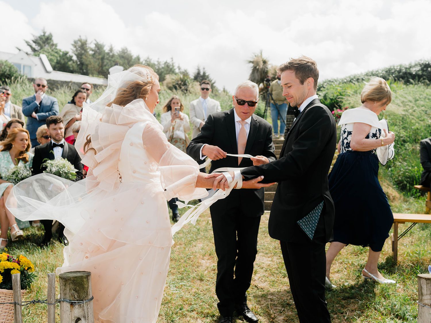 Wedding ceremony taking place outdoors with bride in flowing pink dress and groom in tuxedo.