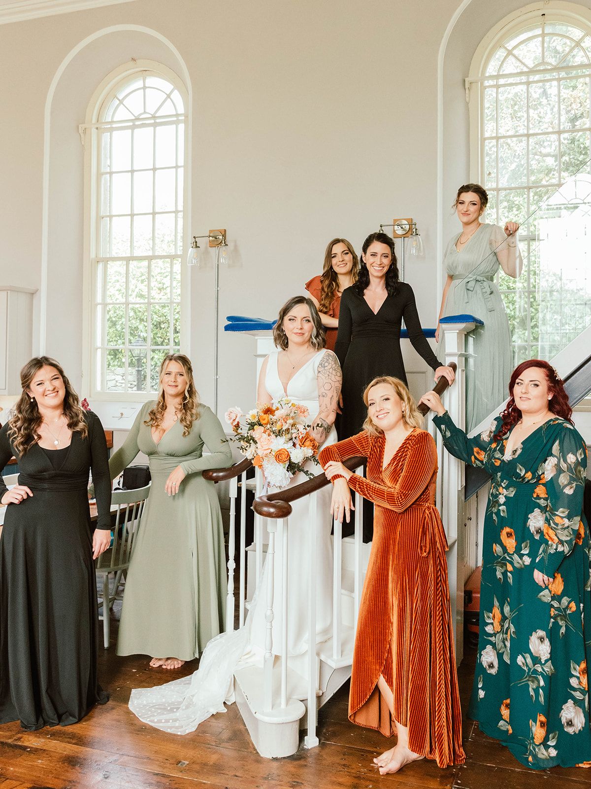 A group of wedding party members in coordinated dresses gather on an elegant white staircase in a bright, sunlit venue.