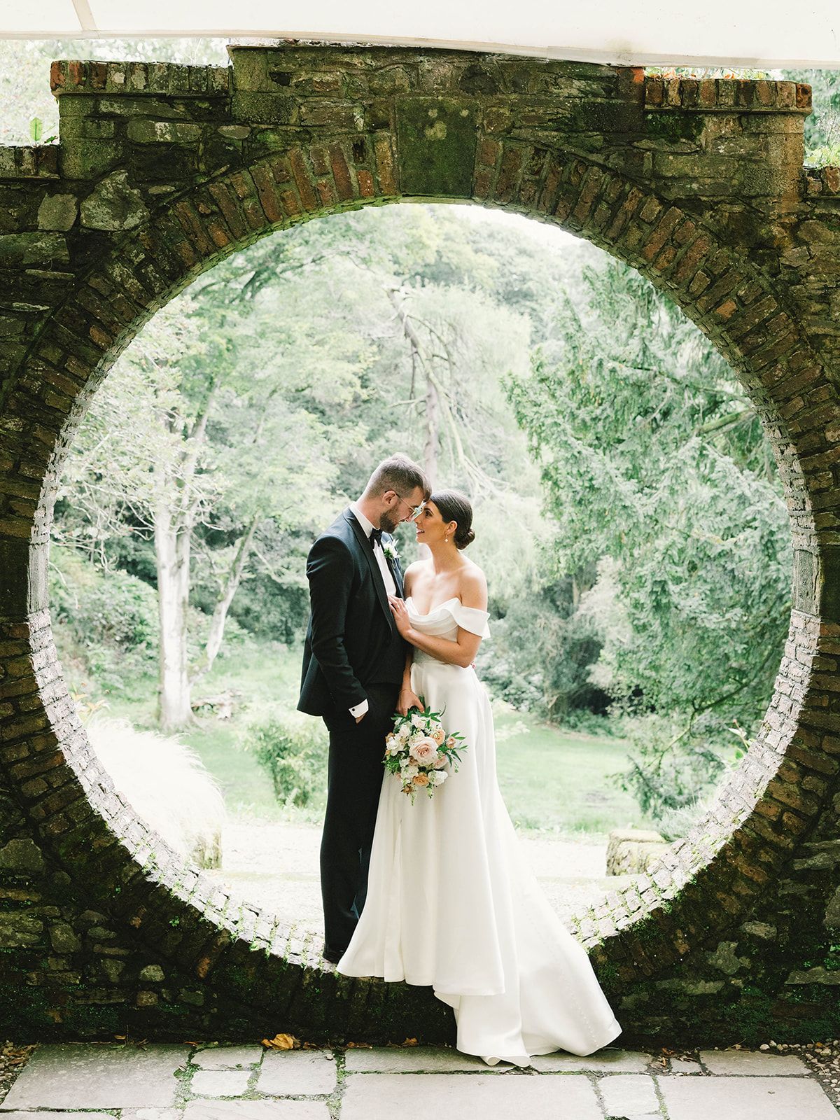A couple shares a romantic moment under a circular stone archway surrounded by lush greenery and trees.