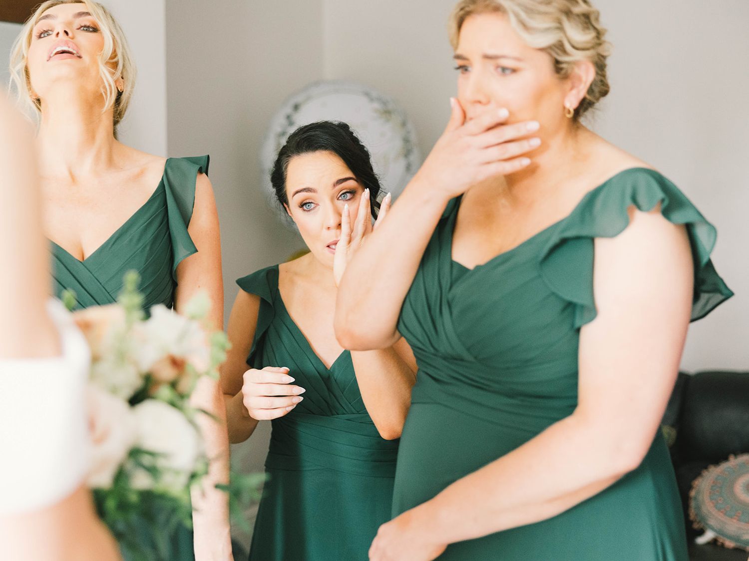 Three bridesmaids in emerald green flutter sleeve dresses share a candid joyful moment at a wedding.