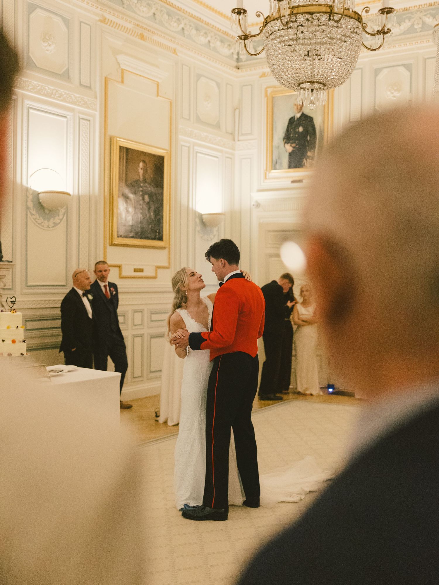 People in formal attire socialize in an elegant ballroom with ornate chandeliers and white decor.
