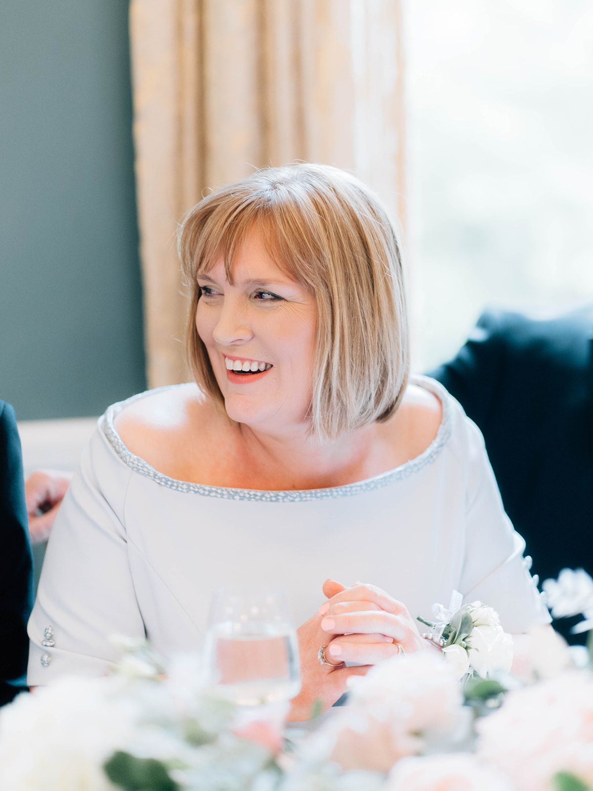 Smiling guest in white dress enjoys reception at wedding with floral table decorations and soft lighting.