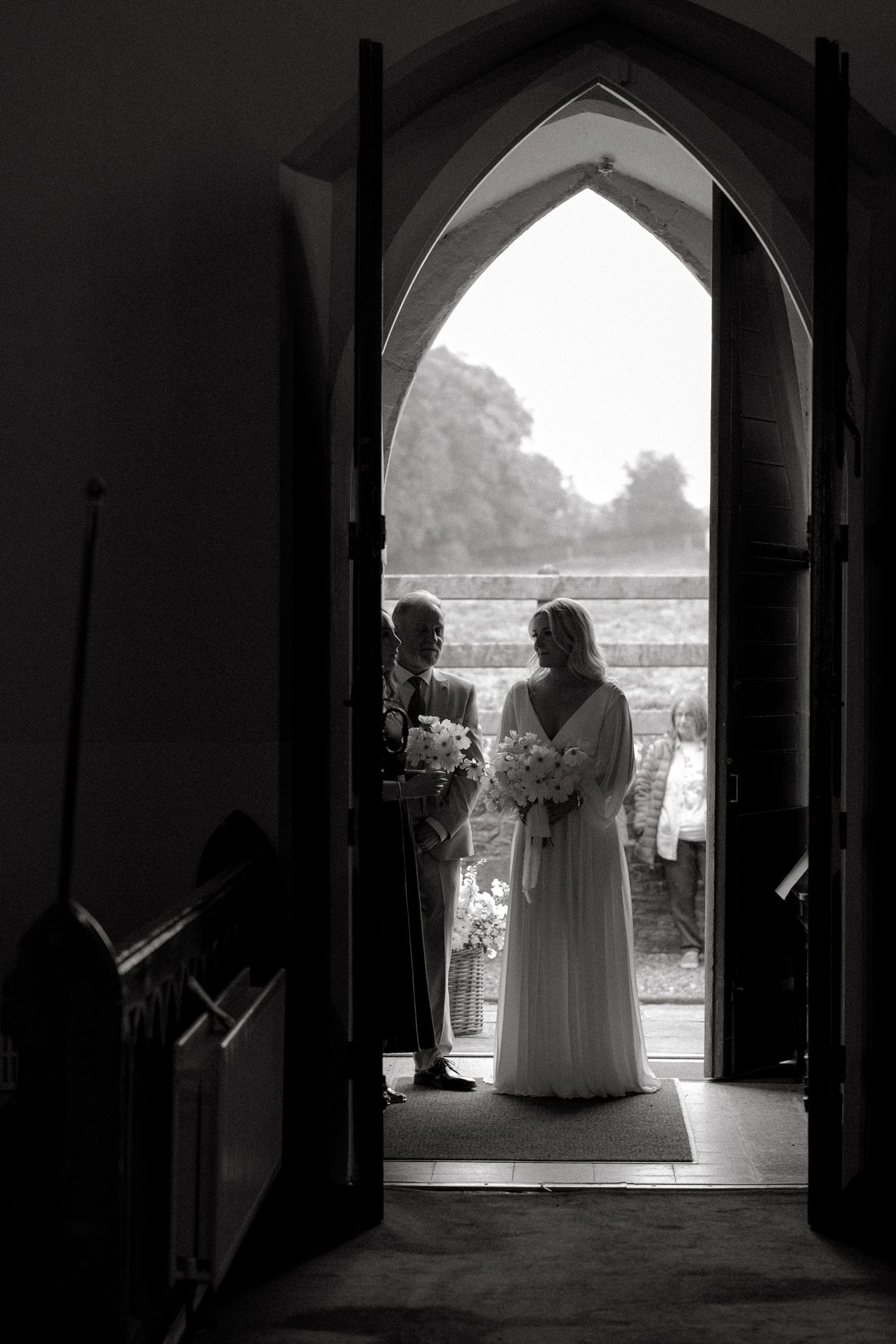 A silhouetted figure stands in a gothic archway doorway creating a dramatic black and white composition.