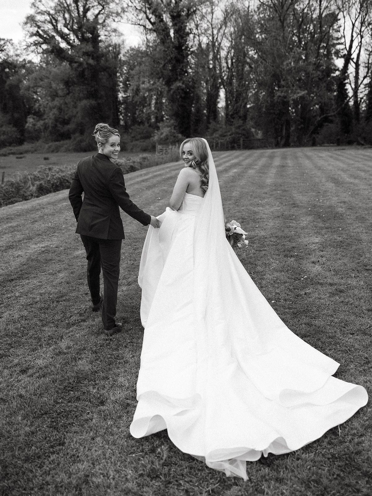 A black and white wedding photo showing a couple walking on a lawn with tall trees in the background.
