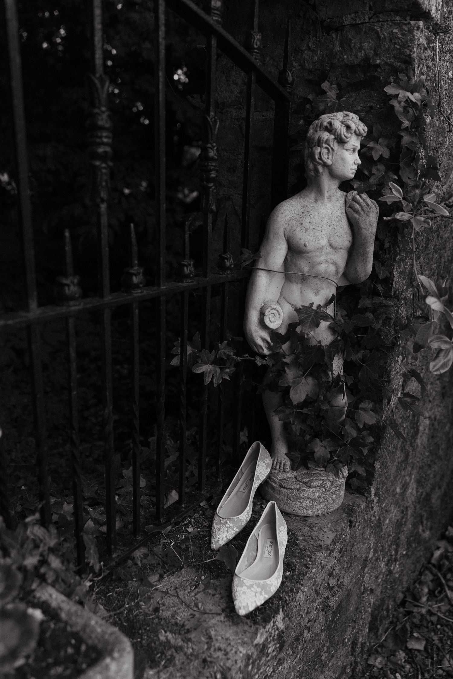 Black and white photo of a classical stone statue beside iron gates, with white ballet flats placed at its base.