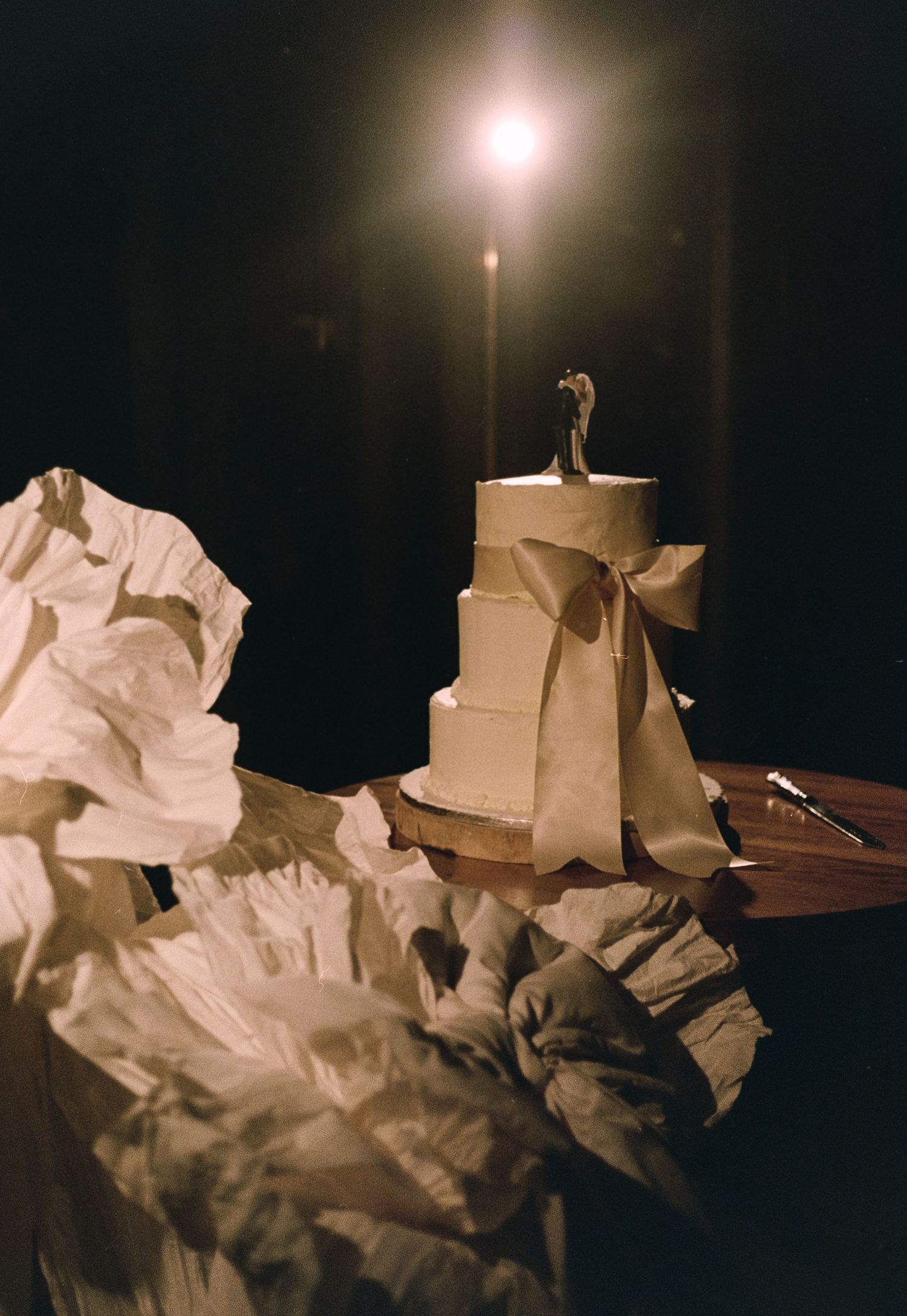 Elegant white wedding cake with bow and bride and groom topper lit by candle in dark romantic setting.