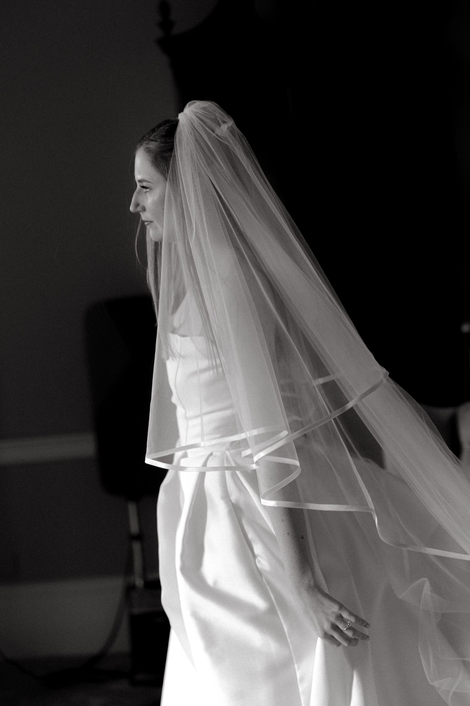 A bride in a white wedding dress and flowing veil stands in dramatic black and white lighting.