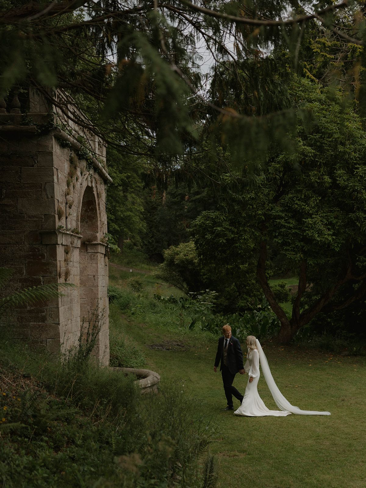 Wedding couple walking through a scenic garden with stone archways and lush greenery.