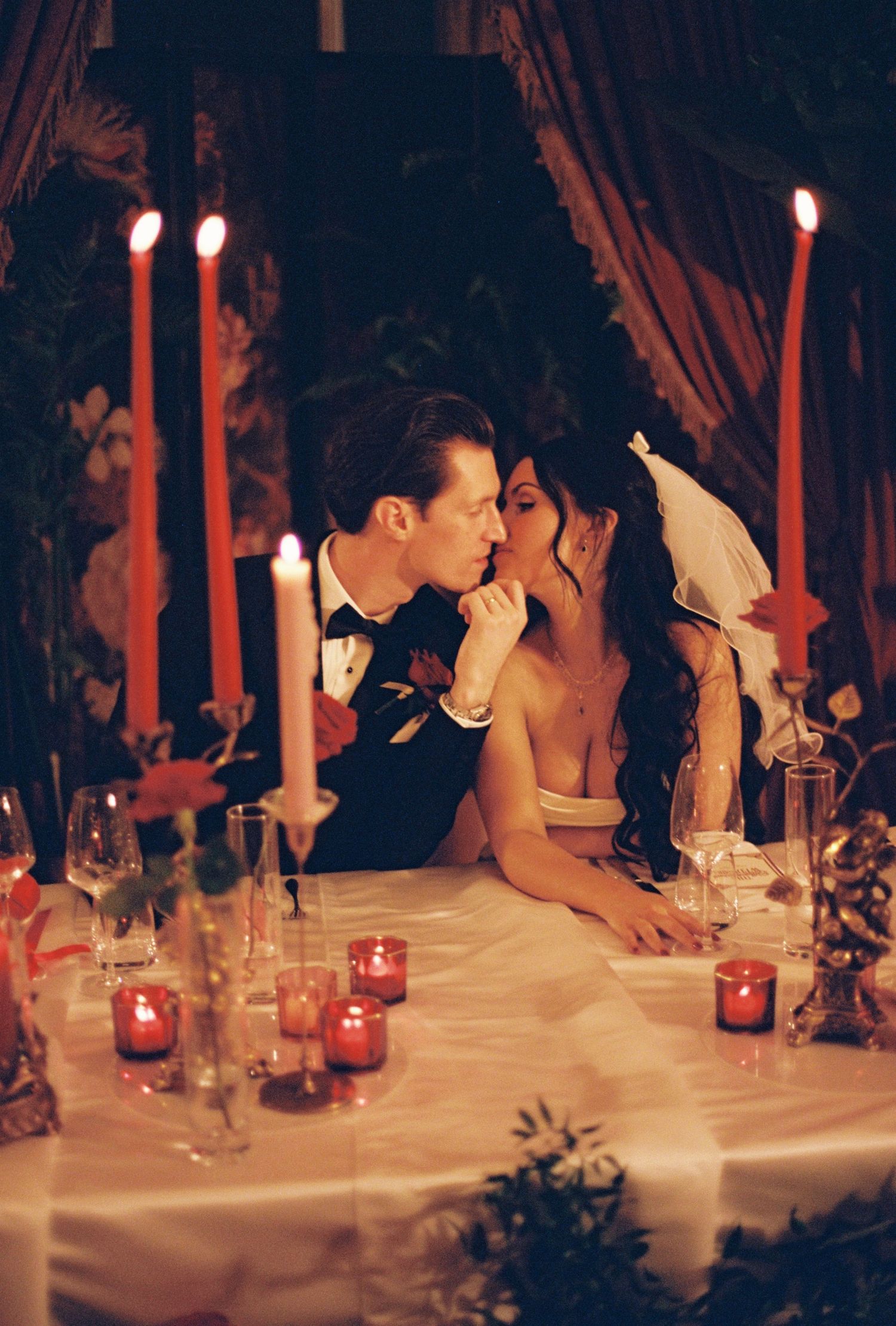 Bride and groom share a romantic kiss at candlelit wedding reception table with red taper candles and warm glowing ambiance.