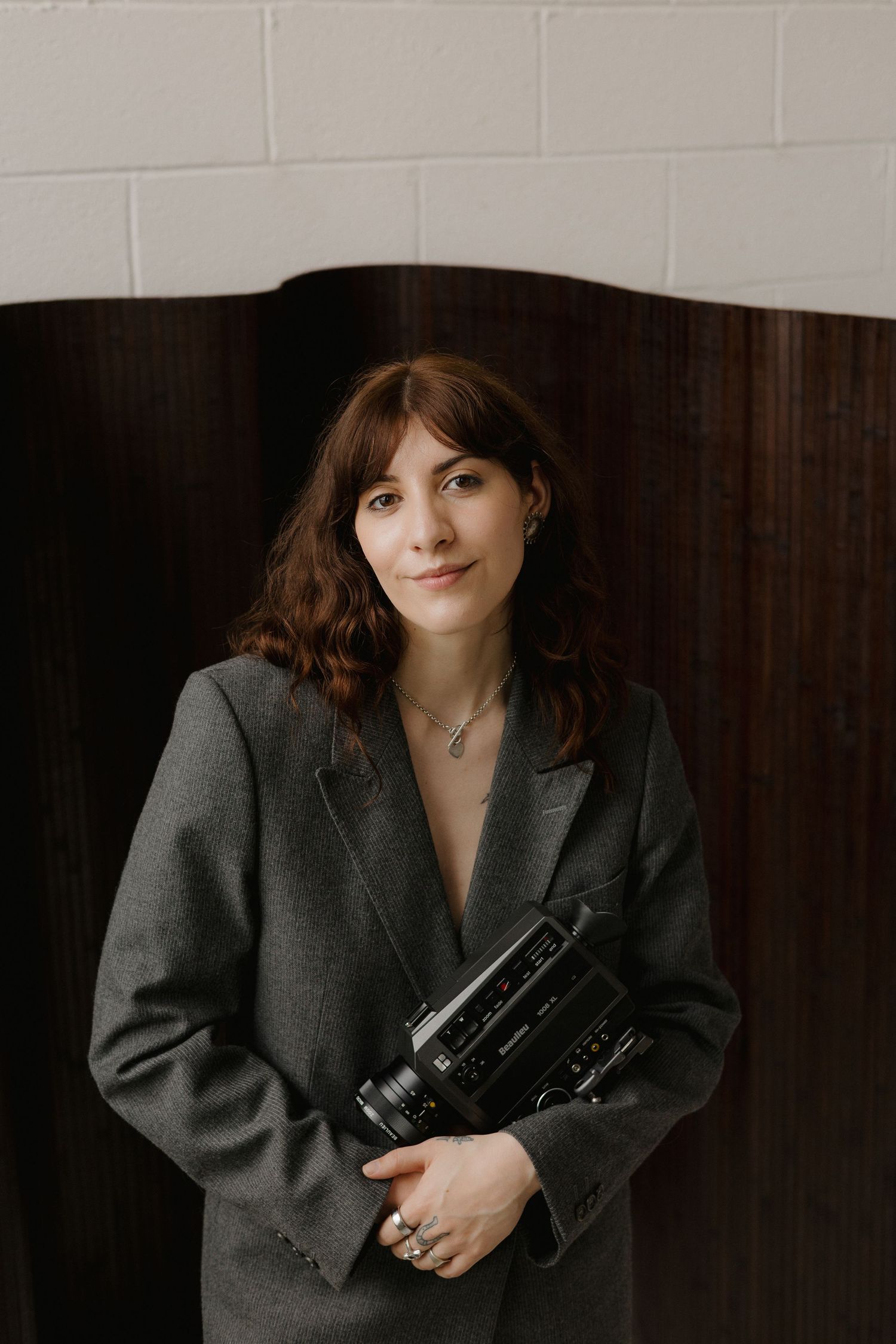 A woman with auburn hair holds a vintage video camera, wearing a gray blazer, posed against a wooden backdrop.
