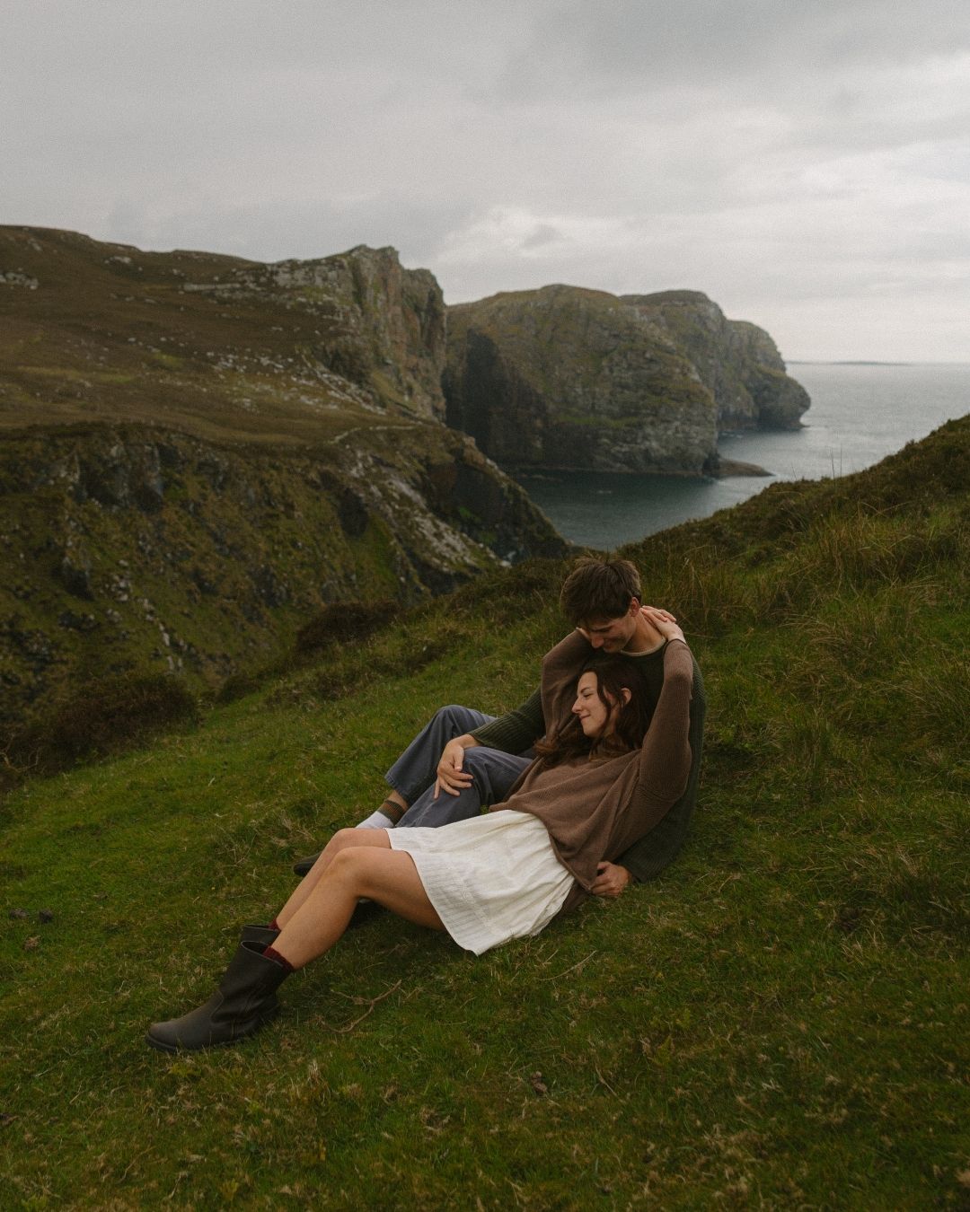 Couple embracing on a lush green cliff overlooking dramatic coastal rock formations under an overcast sky.