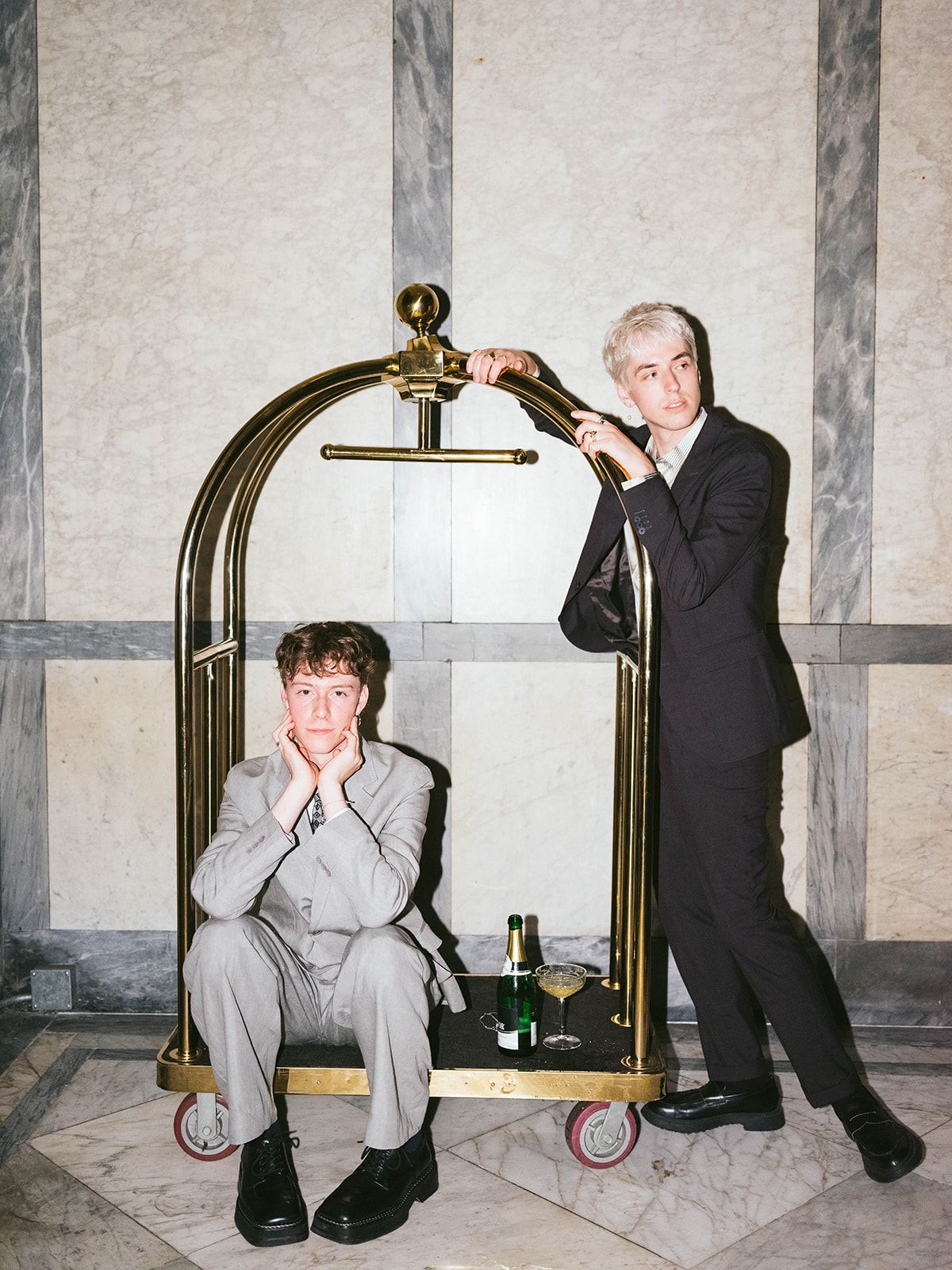 Two men pose playfully with a brass hotel luggage cart in an elegant marble hallway.