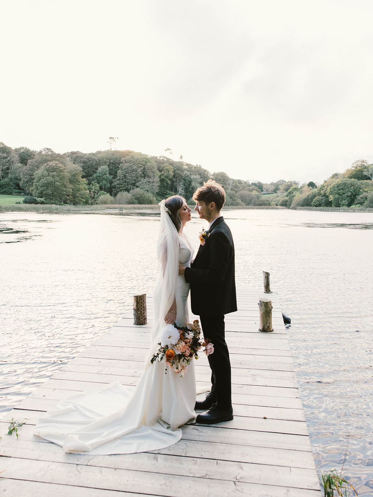 A romantic wedding moment on a wooden dock by the lake with fall foliage in the background.