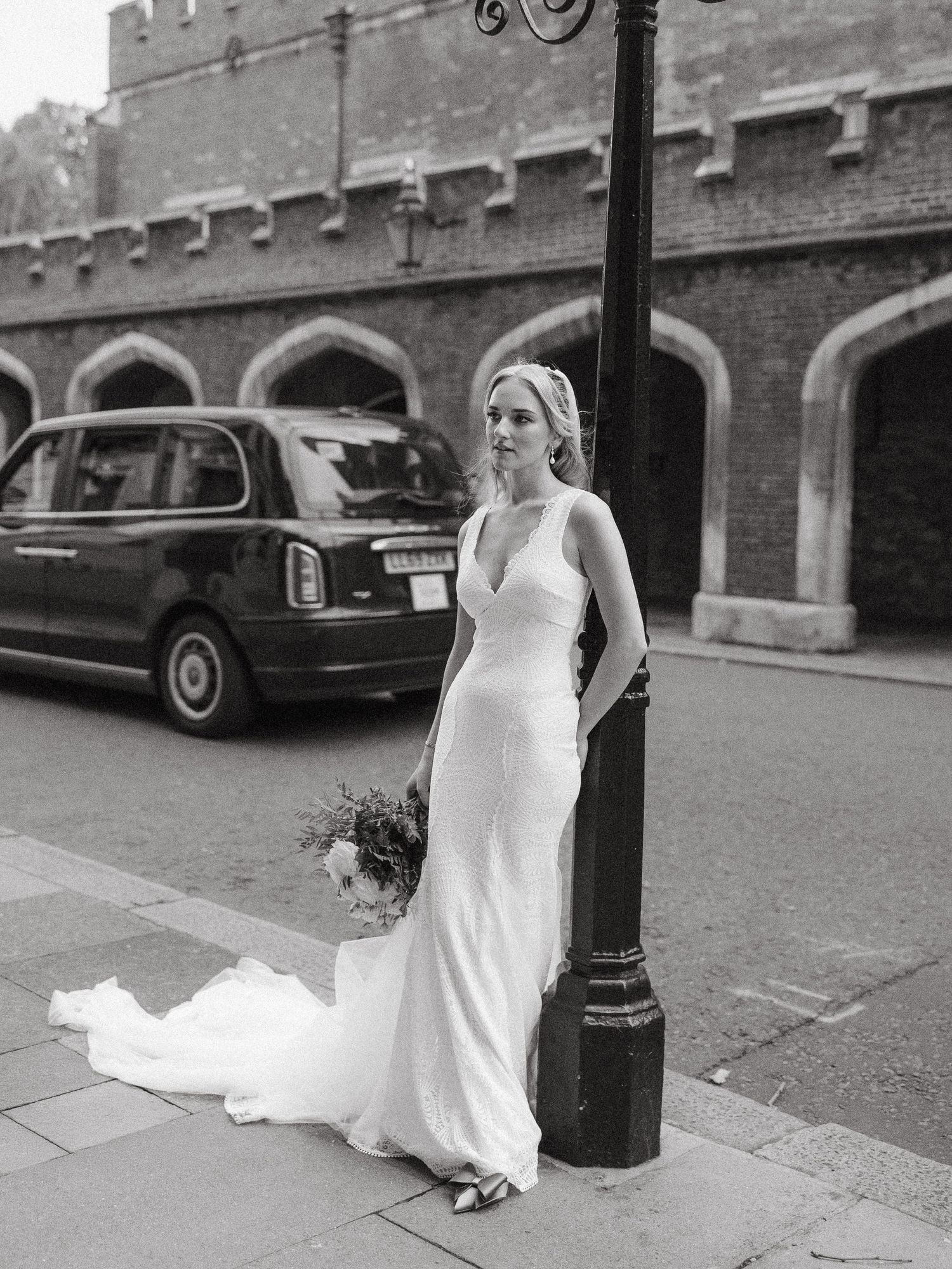 A classic black and white photo of a bride in sleek dress standing beside vintage lamppost near brick building.