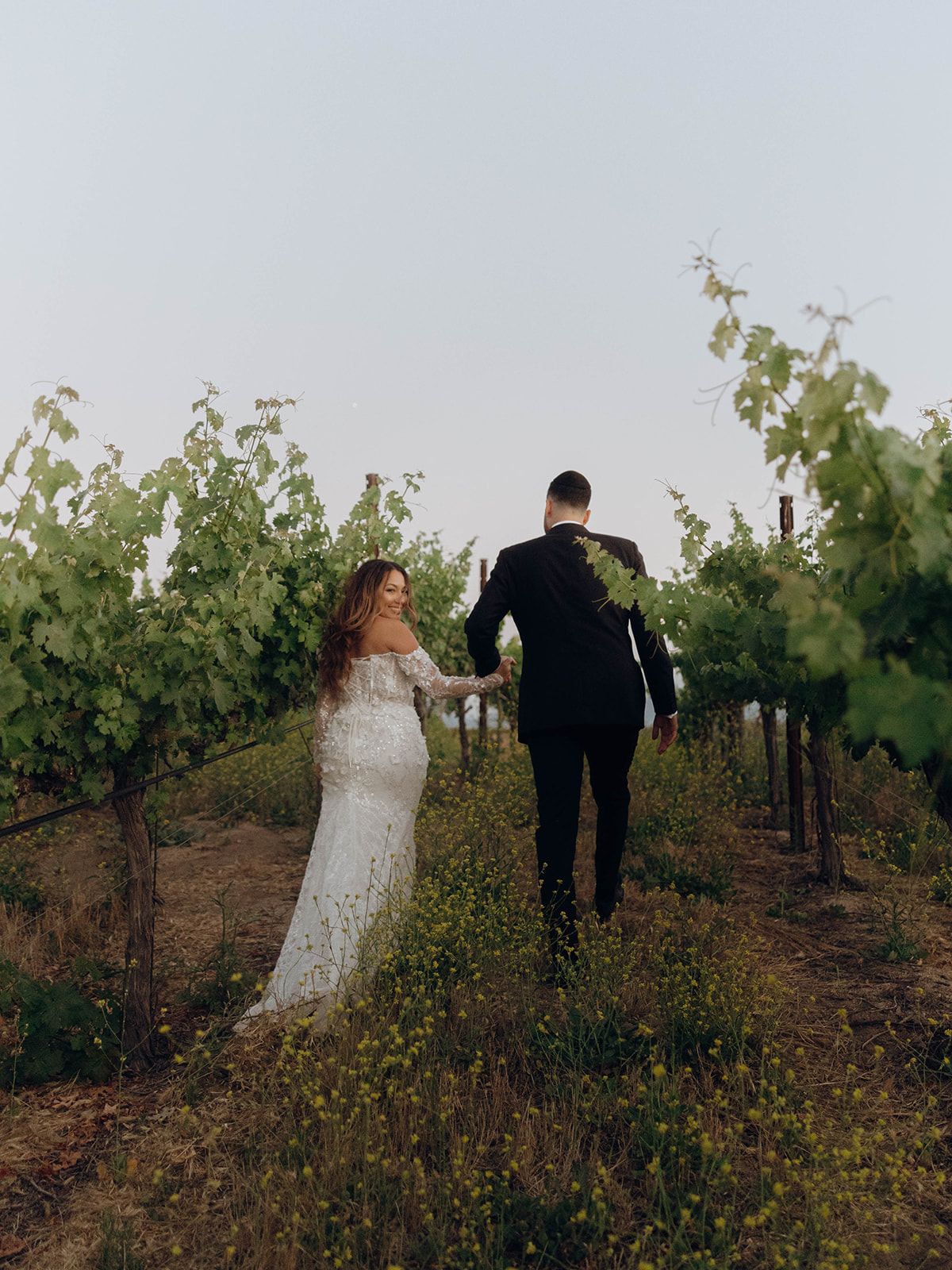 Couple walking through a scenic vineyard at sunset, bride in lace gown and groom in black suit amid grapevines.