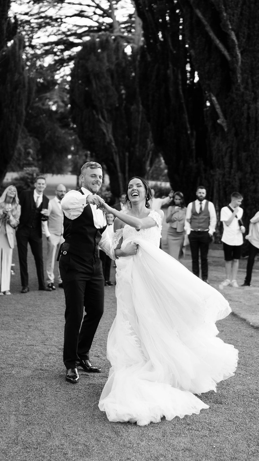 Black and white photo of newlyweds dancing joyfully outdoors surrounded by wedding guests.