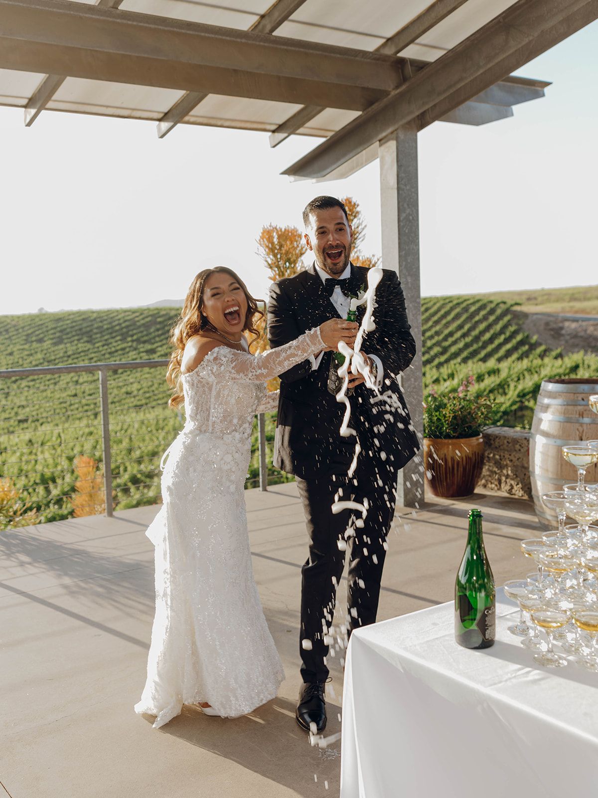 A joyful wedding couple celebrating with champagne on a winery terrace overlooking scenic vineyard rows.