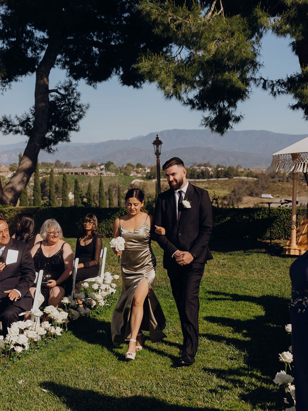 Wedding ceremony outdoors with mountains in background and decorative white floral arrangements on grassy lawn.