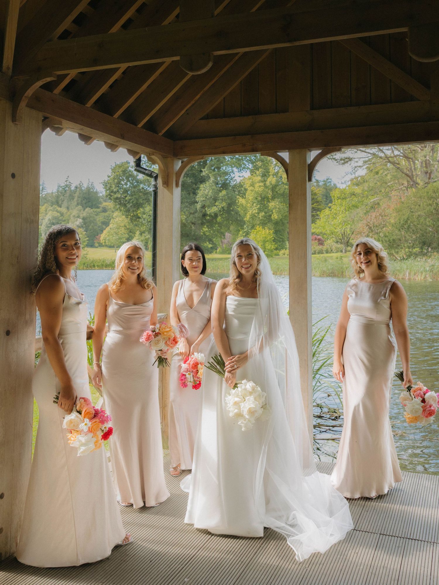Wedding party in white gowns holds colorful bouquets while standing in a waterfront pavilion.
