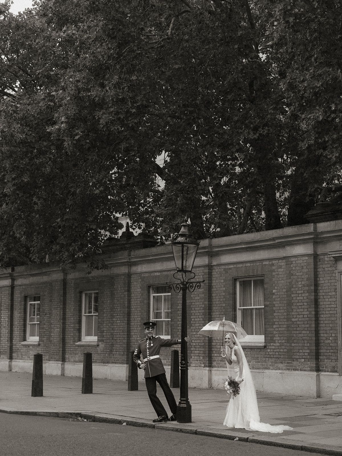 A couple with umbrellas poses in front of a historic brick building with classic lamp posts.