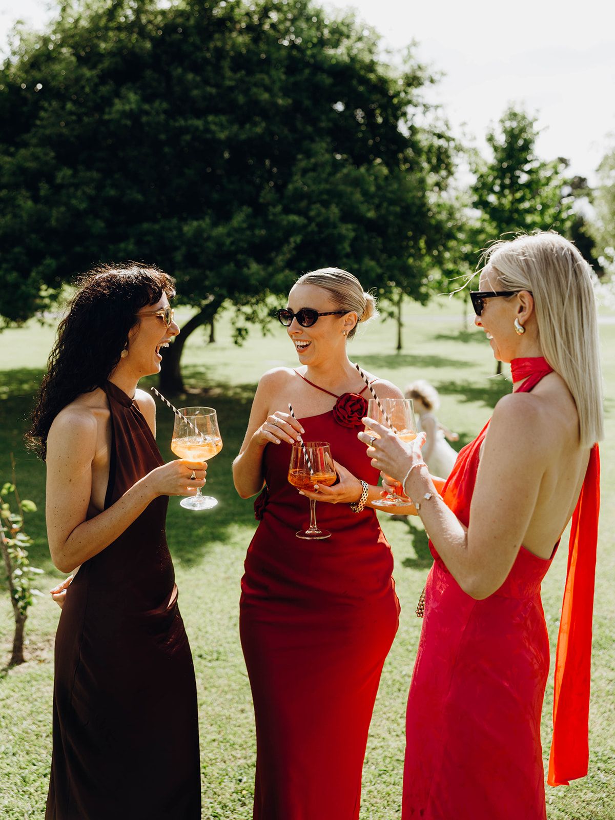 Three friends in elegant dresses toast with wine glasses while enjoying a sunny outdoor gathering.
