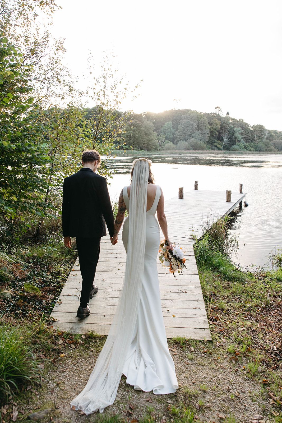 A couple in wedding attire holds hands while strolling down a wooden dock by a serene lake.